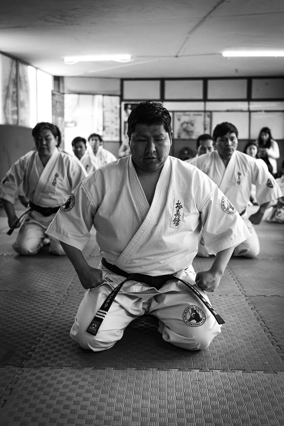 A karate class kneeling in silence on foam tatami, eyes closed, waiting for the first movement.